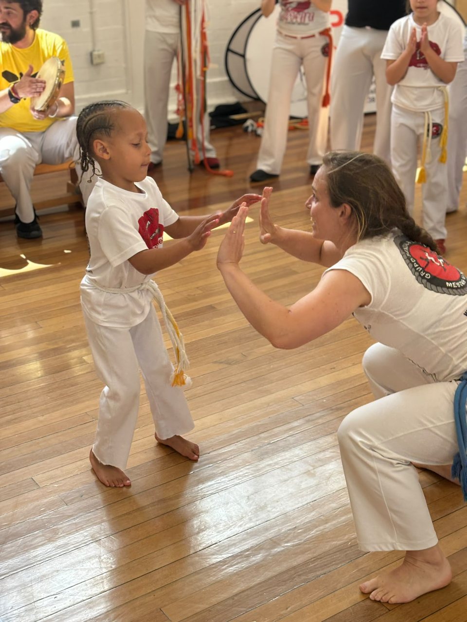 A young capoeirista and an adult crouched at their level share an encouraging moment.