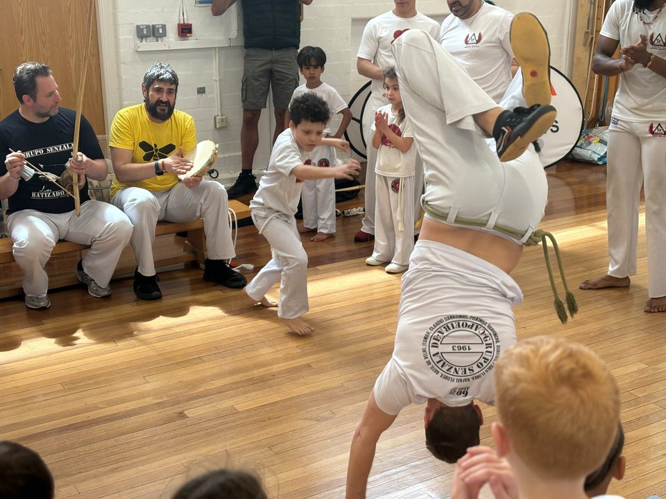 A capoeirista balances upside down in a school roda, with musicians and young students around the circle.